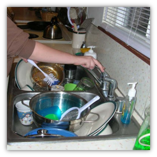 A kitchen sink that is full of dishes, and a person is running the water and holding a brush.