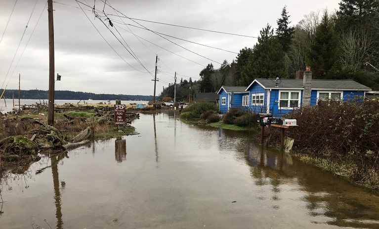 Flooding along the coast of Vashon Island with blue houses on the right and Puget Sound on the left.