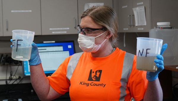 King County staff in orange shirt holding two beakers with water samples