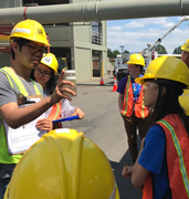 A tour guide holding a container with a sample of treated wastewater for students to view. All people have safety equipment on (hard hats, eye protection and orange vests)
