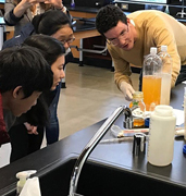 An educator is pointing at a bottle with orange fluid while three high-school students are leaning in to observe. 
